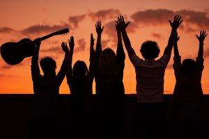 Back view silhouettes of young people with guitar standing with raised arms against colorful sundown sky while having fun and enjoying summer party together